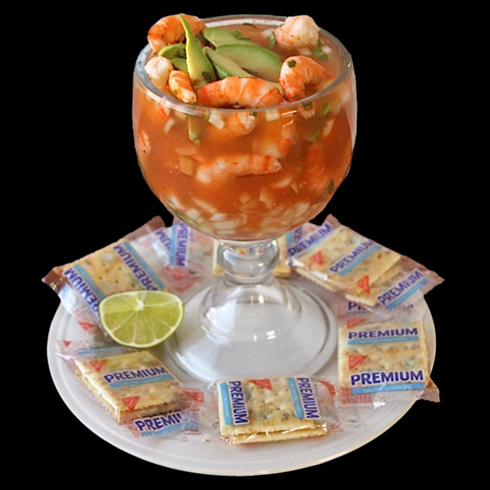 A glass of shrimp cocktail with avocado and lime, surrounded by cracker packets on a white plate.