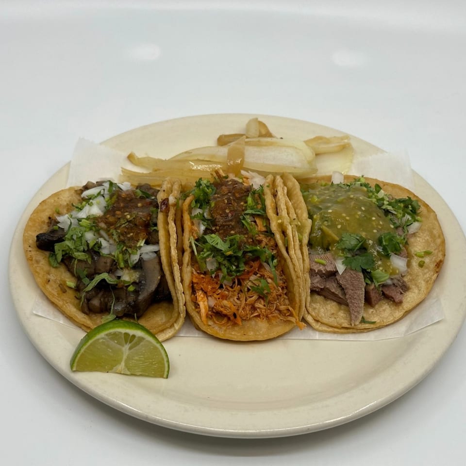 Three Taqueria Mi Pueblo tacos with various fillings, onions, cilantro, and salsa, with a lime wedge.