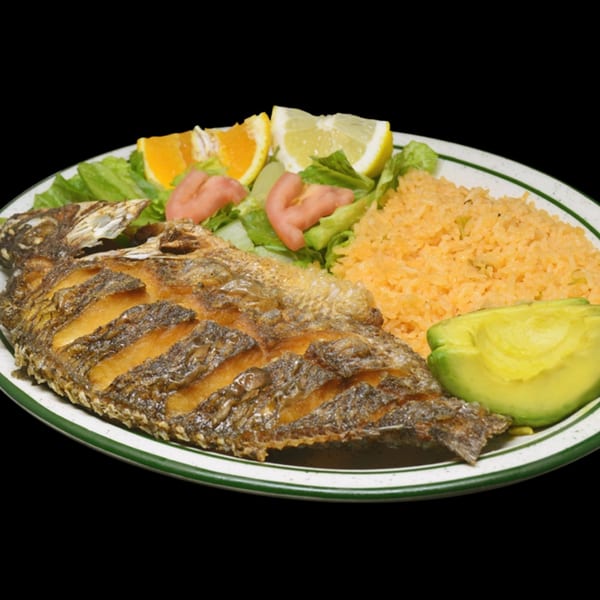 Whole fried fish with rice, salad, avocado, and citrus slices on a white plate with green rim.