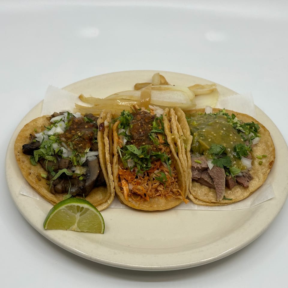 Three Taqueria Mi Pueblo tacos with various fillings, onions, cilantro, and salsa, with a lime wedge.